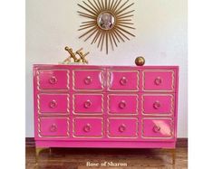 a pink dresser with gold trim and knobs on the doors is shown in front of a white wall