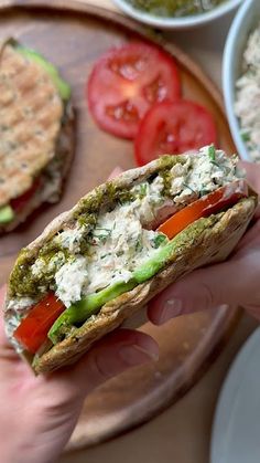 a hand holding a sandwich with tomatoes, cucumber and other food items in bowls