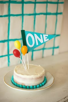 a birthday cake decorated with balloons and a one flag on top is sitting on a plate