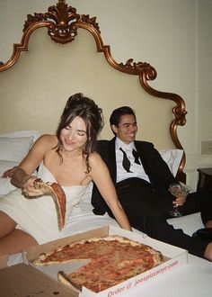 a man and woman sitting on a bed with pizza in front of them