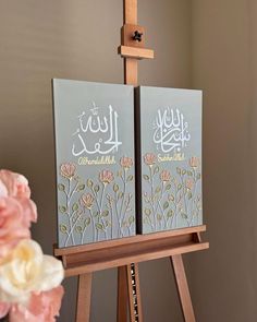 two paintings on easel with flowers in vase and wall behind them, one has arabic calligraphy