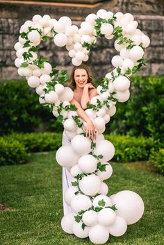 a woman standing in front of a heart shaped balloon arch with greenery on it