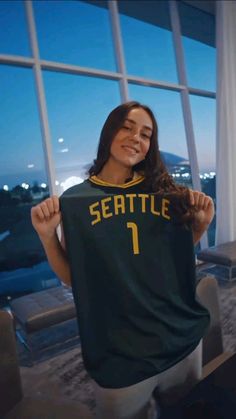 a woman holding up a seattle basketball jersey in front of a large window at night