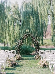 an outdoor ceremony setup with white chairs and flowers on the aisle, surrounded by willow trees