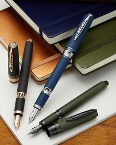 three fountain pens sitting next to each other on top of a table with notebooks