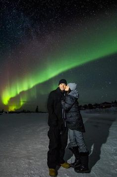 two people are standing in the snow with an aurora light behind them and one person is kissing