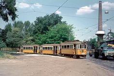 an old yellow train traveling down tracks next to a green truck and power line with trees in the background