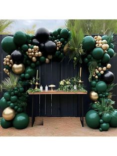 a table topped with lots of balloons and greenery next to a black fence covered in gold balls