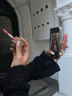 a woman taking a selfie with her cell phone and some lipstick in front of a building