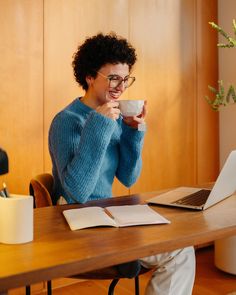 a woman sitting at a table with a laptop and cup in her hand, drinking coffee