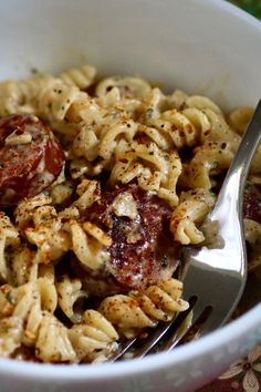 a white bowl filled with pasta and meat covered in sauce next to a fork on top of a table