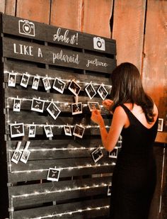 a woman in a black dress writing on a wooden board with polaroid pictures attached to it