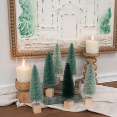 small christmas trees sit on blocks in front of a painting and candle holder with two lit candles