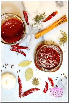 two jars filled with red sauce next to some spices and herbs on a marble surface