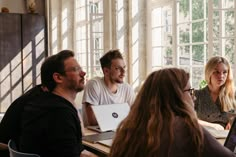 four people sitting at a table with laptops and papers in front of them, all looking at each other