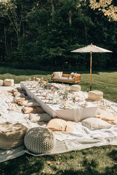 an outdoor table set up with white linens, pillows and food on the ground