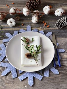 a white plate topped with a napkin covered in pine cones and greenery on top of a wooden table