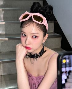 a woman wearing pink sunglasses and a black choker is sitting on stairs with her hand under her chin