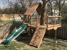 a wooden play structure with a slide and climbing wall in the back yard next to a tree