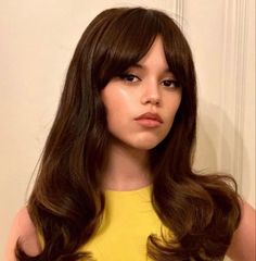 a woman with long brown hair wearing a yellow top and posing for the camera in front of a white wall