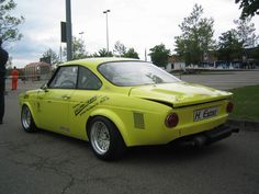 a yellow car parked on the side of a road next to a parking lot with people standing around