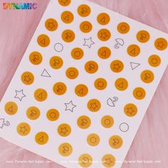 A sheet of nail art stickers features orange circles adorned with black question marks, capturing the enigmatic essence of Squid Game. Black outlined shapes such as triangles, stars, and crescent moons add an element of intrigue against a gentle pink background. The Dynamic Nail Supply brand name is prominently displayed at the bottom. Squid Game Marble, Squid Game, Squid Games, Nail Supply