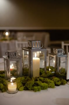 candles and moss are on the table with white linens, which is decorated with silver lanterns