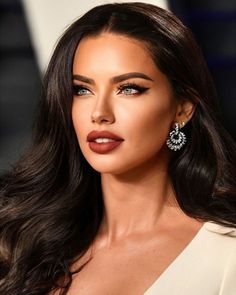 a woman with long dark hair wearing earrings and a white dress is looking at the camera