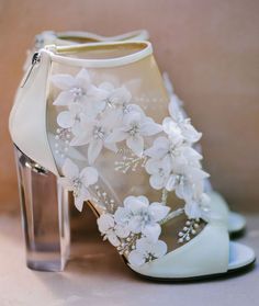 a pair of white high heel shoes with flowers on the side and clear heels in front