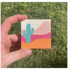 a hand is holding up a small square painted with cactus and pink, orange, and blue colors