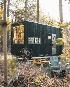 a small black cabin in the woods with two chairs and a picnic table set up outside