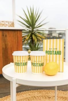 two yellow cups sitting on top of a white table next to a lemon and a postcard