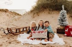 three children are sitting on the beach holding a sign that says happy new year and christmas trees