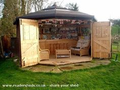 a wooden shed with two chairs and a bar in the back yard, surrounded by green grass