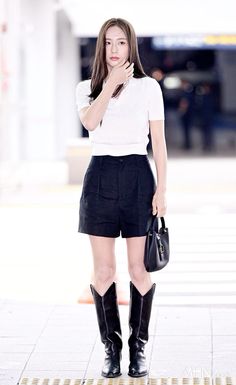 a woman in black shorts and white shirt standing on the sidewalk with her hand under her chin