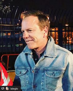 a man wearing a jean jacket standing in front of a fence