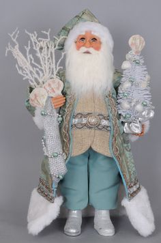 a santa clause doll holding two christmas trees and ornaments in his hands, on a gray background