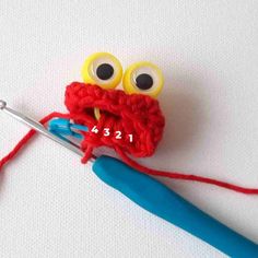 a crocheted red object with eyes and a pair of blue handled knitting needles