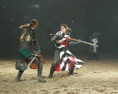 two people dressed in medieval costumes playing with swords and shieldes on a dirt field