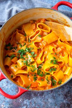 a red pot filled with pasta and sauce on top of a blue table cloth next to a wooden spoon