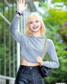 a woman with blonde hair wearing black leather pants and a grey crop top waving to the camera