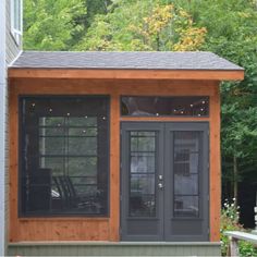 a small wooden building with glass doors and lights on the windows, in front of some trees