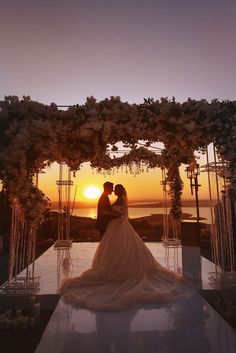 the bride and groom are standing under an arch with flowers on it at sunset in front of the ocean