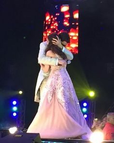 a man and woman embracing on stage at a wedding ceremony with bright lights in the background