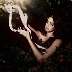 a woman is holding a snake in her hand and posing for the camera with flowers around her