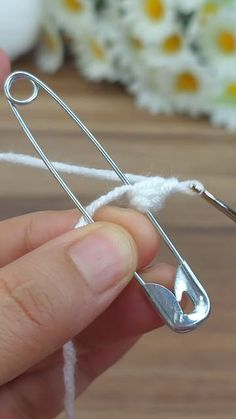 a person is holding a piece of white yarn with scissors in the foreground and flowers in the background