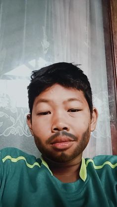 a man with a mustache and green shirt looking at the camera while sitting in front of a window