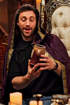 a man dressed in medieval garb holding a cup and looking at the camera while sitting on a chair