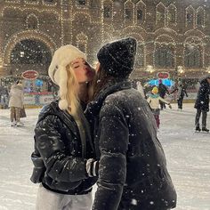 two people standing in the snow kissing each other and wearing winter clothes with lights behind them