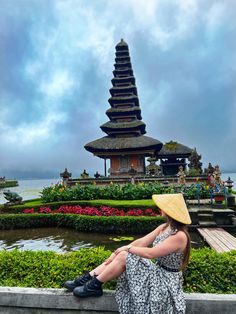 a woman in a hat sitting on a wall looking at the water and pagodas